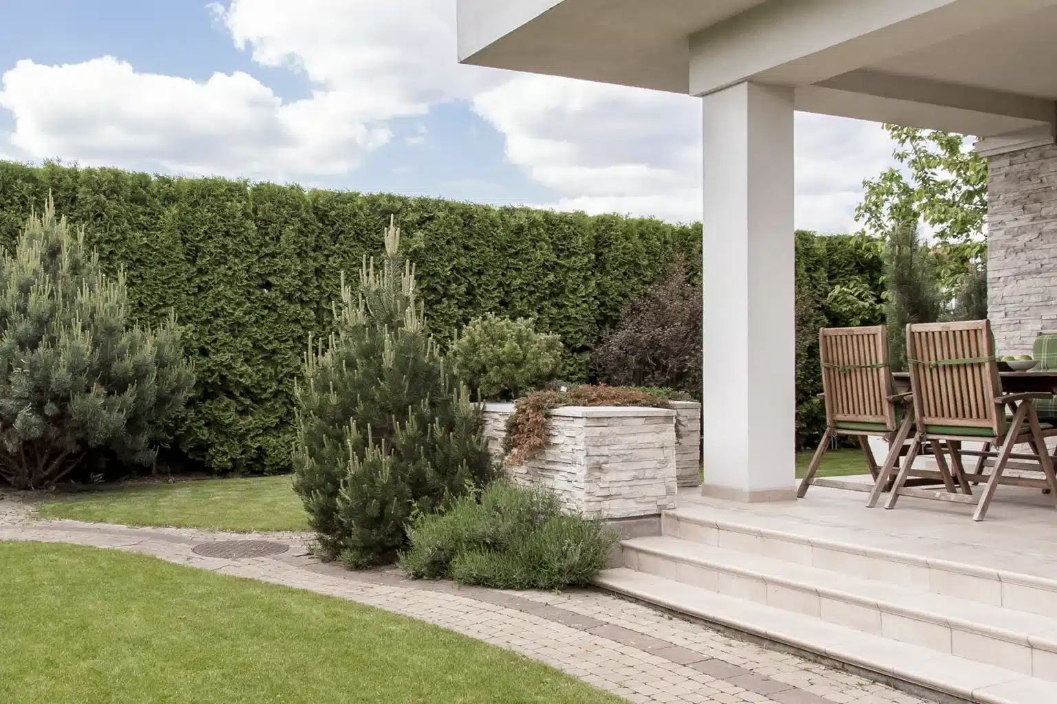 A private terrace featuring a dining area with wooden chairs and a table, surrounded by lush greenery. The terrace is elevated, with stone steps leading down to a well-manicured lawn. The garden includes neatly trimmed shrubs and tall conifer trees, providing a natural privacy screen. The sky is partly cloudy, adding to the serene and well-maintained outdoor space.