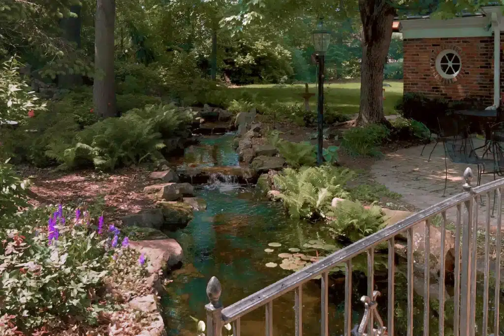 A tranquil garden scene featuring a small stream with lily pads, bordered by rocks and lush greenery, including ferns and blooming flowers. A stone patio with wrought iron furniture is visible near a large tree. In the background, there is a brick building with a circular window. The setting is shaded by trees, creating a peaceful and inviting outdoor space.