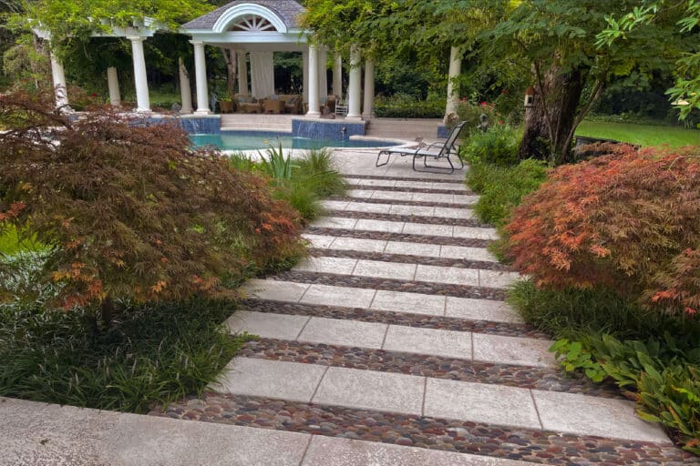 A picturesque backyard scene featuring a stone staircase flanked by lush greenery and Japanese maples, leading down to an elegant white gazebo and a pool. The gazebo houses comfortable lounge furniture, all set in a tranquil garden atmosphere.