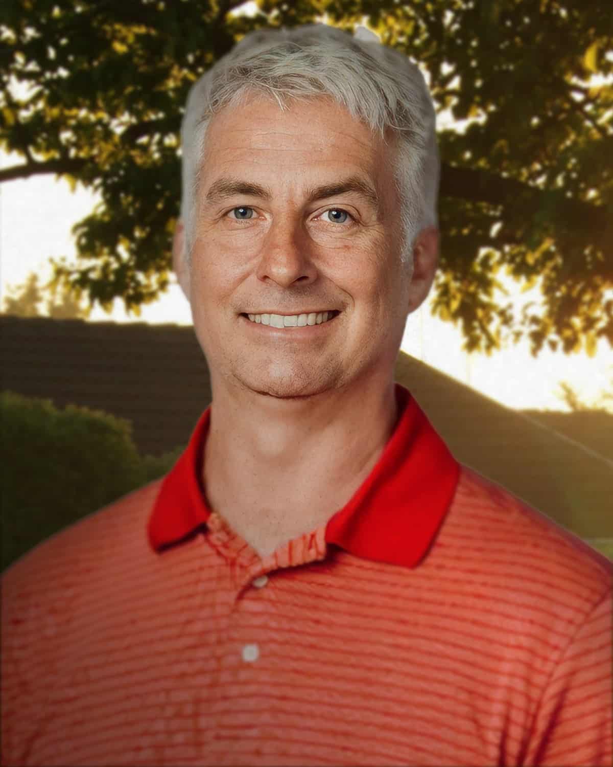 A portrait of a smiling man with short, gray hair wearing a red collared shirt with a striped pattern. The background features a softly focused outdoor scene with trees and warm, golden light filtering through the leaves, suggesting a pleasant evening setting.