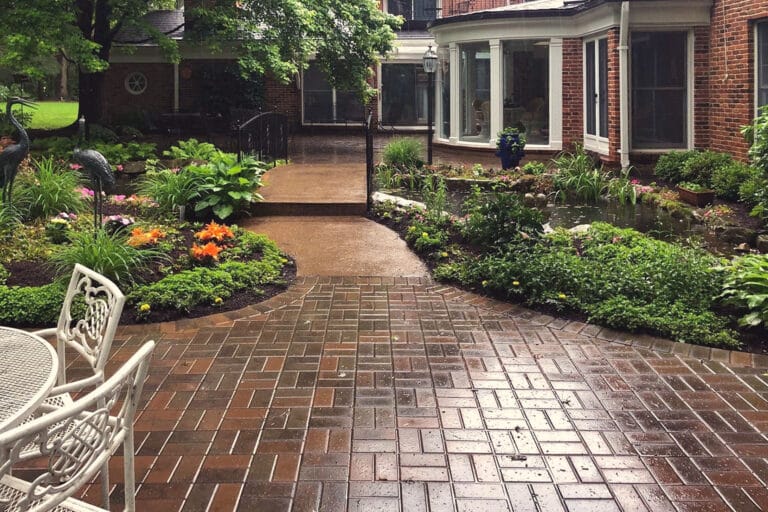 A serene, rain-kissed courtyard featuring a brick paver patio with various seating areas surrounded by lush, vibrant gardens. The curved pathways lead towards a traditional brick house with bay windows, enveloped by a variety of colorful flowers and green shrubbery. A quaint garden pond, visible in the background, adds a touch of tranquility to the scene.