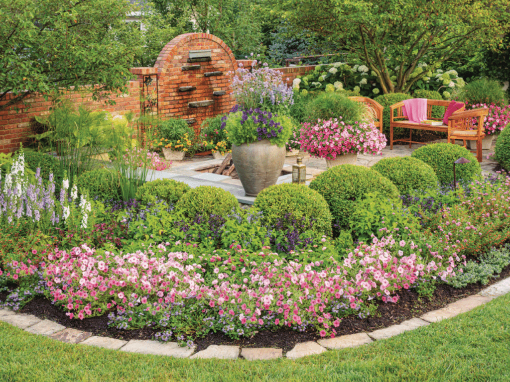 Lush romantic garden featuring boxwood spheres, pink petunias, and purple flowers around a large stone urn, with brick wall and wooden bench