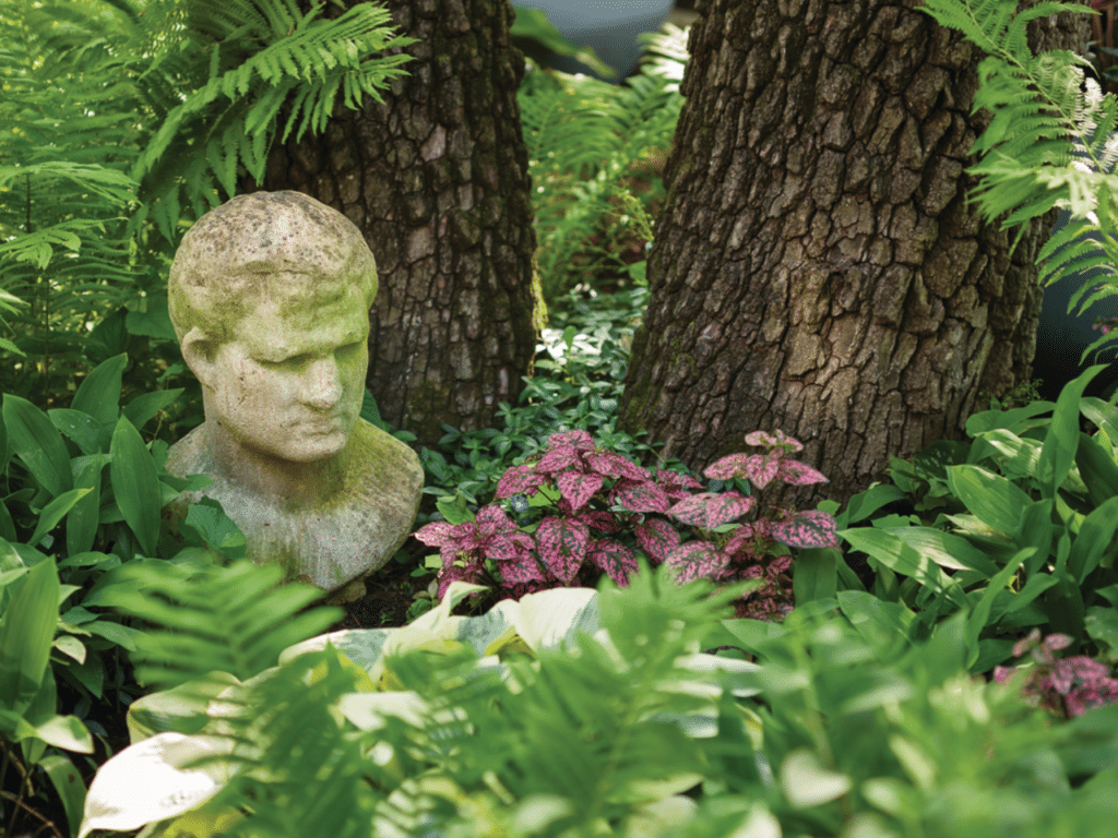 Classical stone bust statue nestled among lush ferns and pink-speckled foliage creating a romantic garden setting