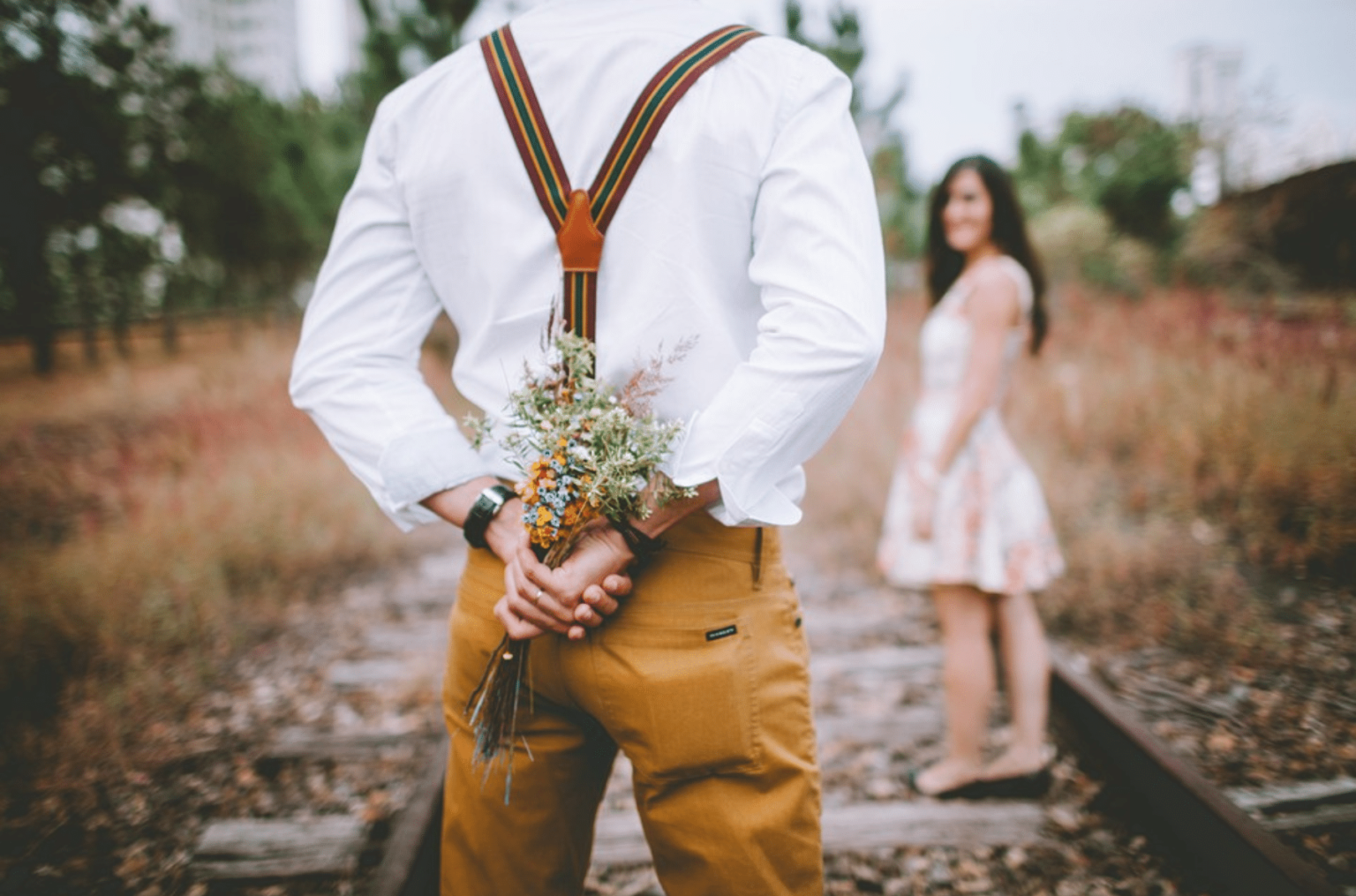 Person in white shirt and suspenders holding wildflower bouquet behind back, with someone in a dress visible in background