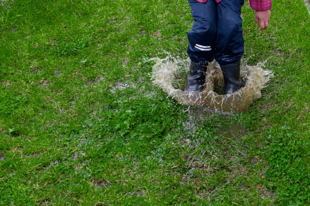 Child in black rubber boots jumping into a puddle in a grassy yard, illustrating standing water and the need for a rain garden in St. Louis.