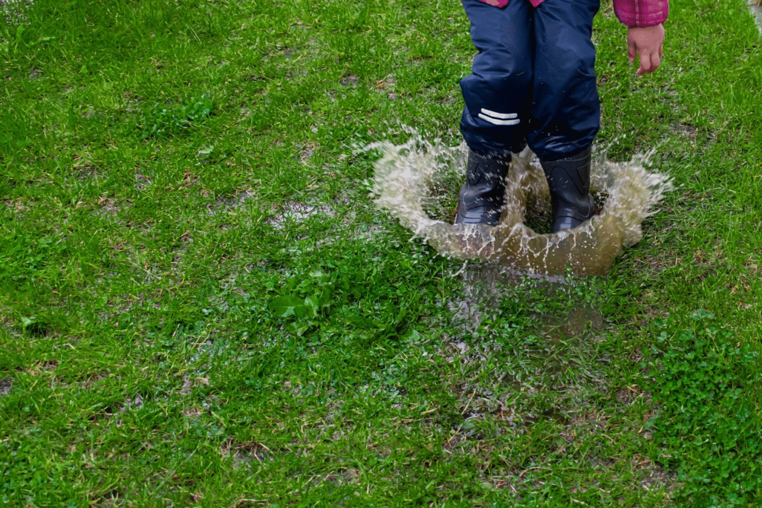Child in black rubber boots jumping into a puddle in a grassy yard, illustrating standing water and the need for a rain garden in St. Louis.