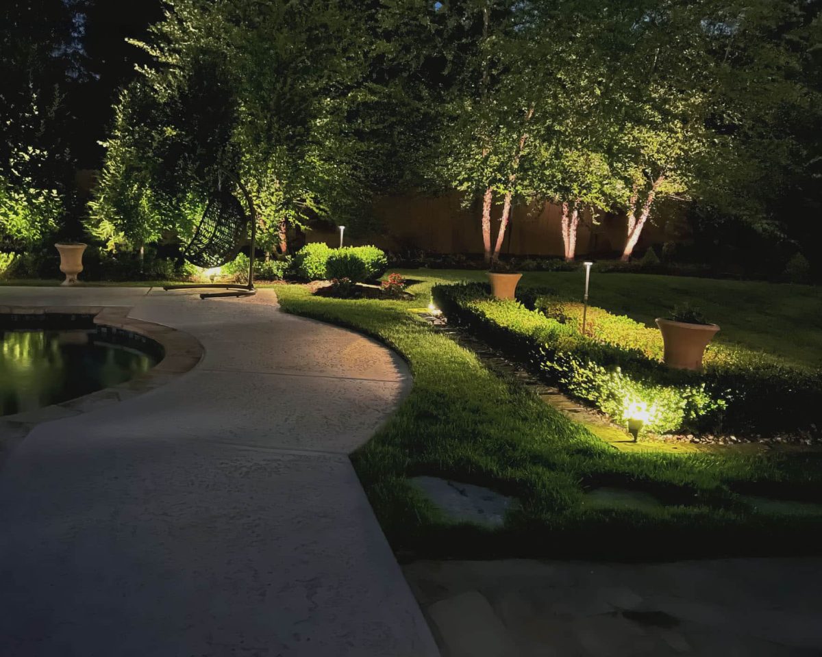 Nighttime view of a serene garden with a curved pathway lit by soft landscape lighting, showcasing a swing, manicured bushes, and reflective pool under the gentle glow of tree lights.