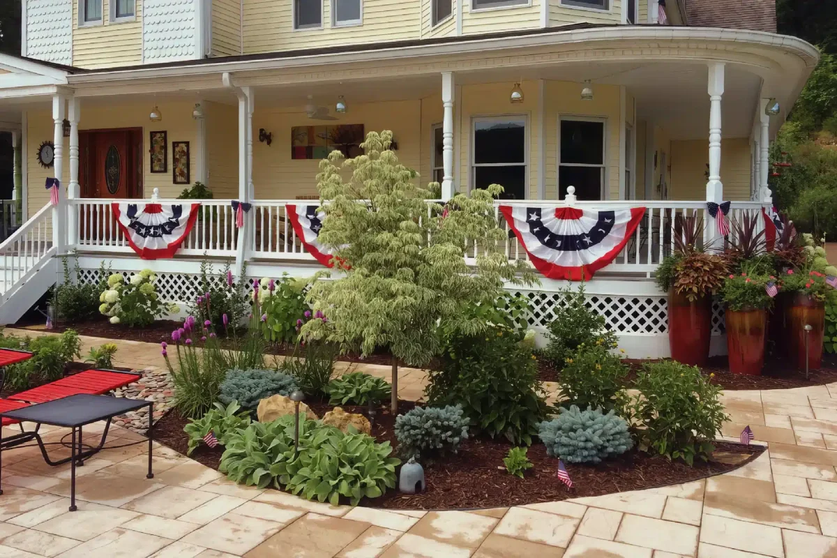 A beautifully landscaped front yard with a paved walkway leading to a yellow house with a large porch. The porch is decorated with patriotic red, white, and blue bunting. The garden in front of the porch features a variety of plants, including a small tree, lush greenery, and blooming flowers. Red lounge chairs are placed on the paved area, enhancing the inviting and festive atmosphere of the scene.