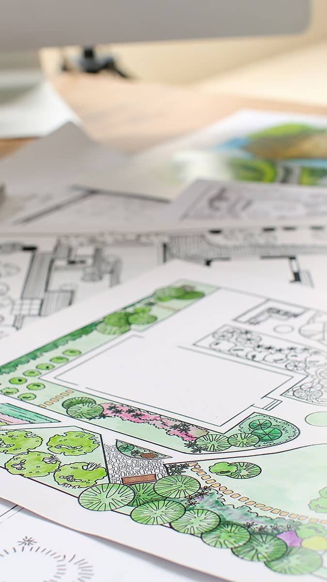 A close-up of detailed landscape design plans on a desk. The plans feature colored sketches of a garden layout, including pathways, trees, and various plants. The background shows a blurred computer monitor and other design sheets, indicating a workspace dedicated to landscape architecture.