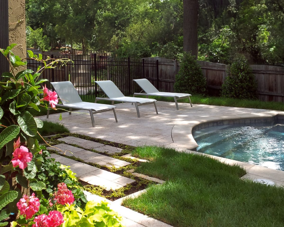 A relaxing poolside setting in a private backyard featuring three white lounge chairs on a stone patio adjacent to a curving swimming pool. The area is lush with vibrant flowers and greenery, partially shaded by mature trees, creating a tranquil oasis.