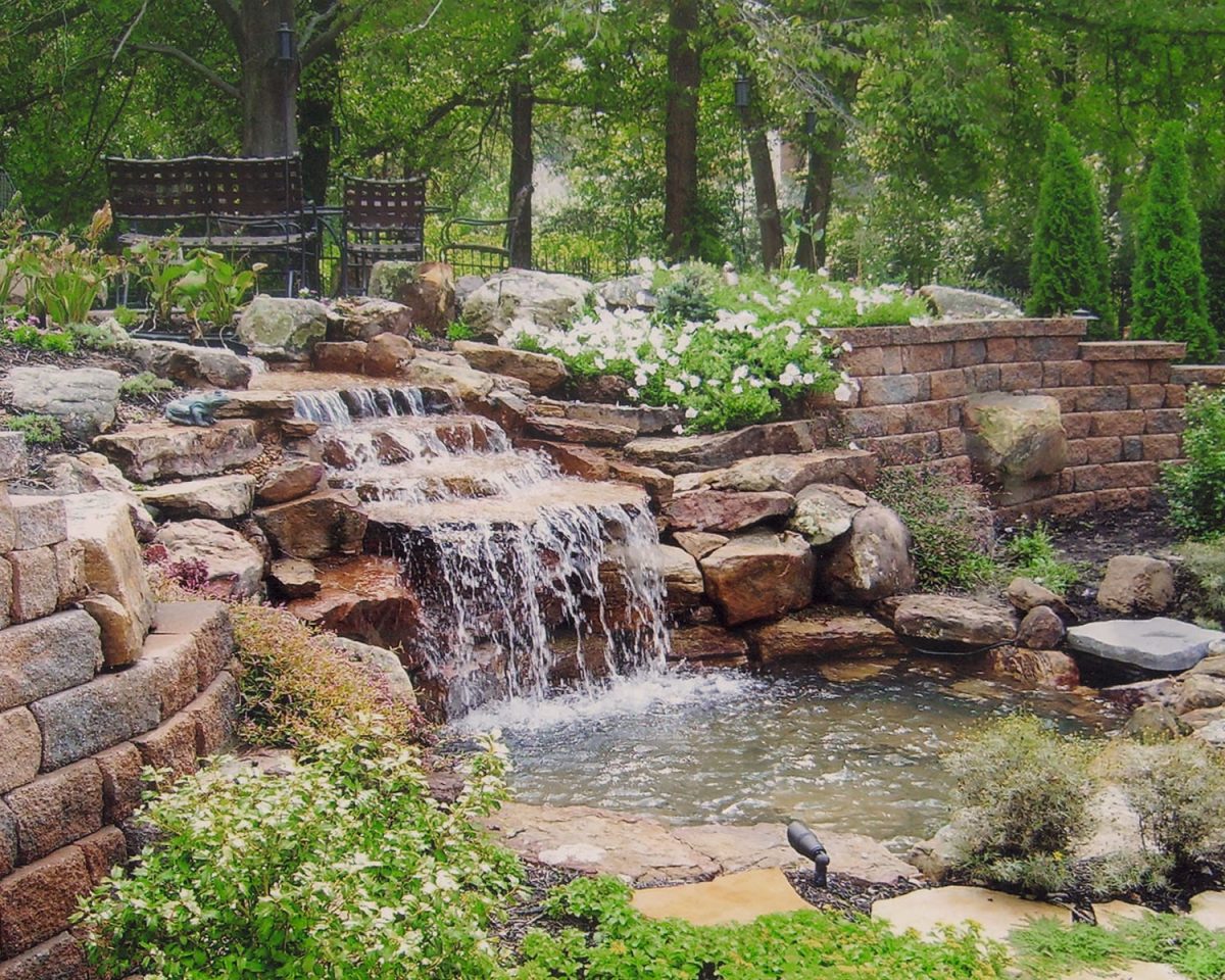 A naturalistic garden waterfall cascades over rocky tiers into a small pond, flanked by terraced stone retaining walls adorned with diverse plantings. Above the falls, a secluded sitting area with a dark iron bench offers a serene viewing spot, surrounded by lush greenery and flowering plants.