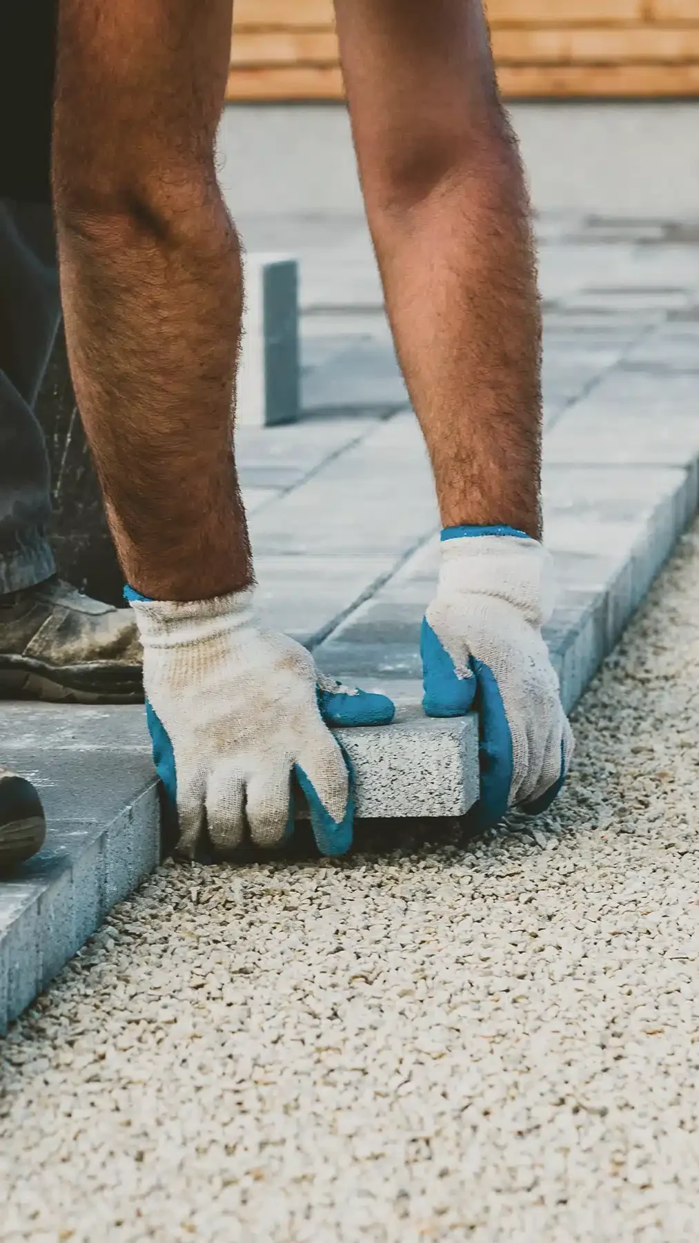 A close-up of a person’s hands wearing white and blue work gloves, placing a concrete paver into position on a gravel bed. The image shows part of the person’s legs and work boots, emphasizing the construction and hands-on aspect of laying a stone patio.