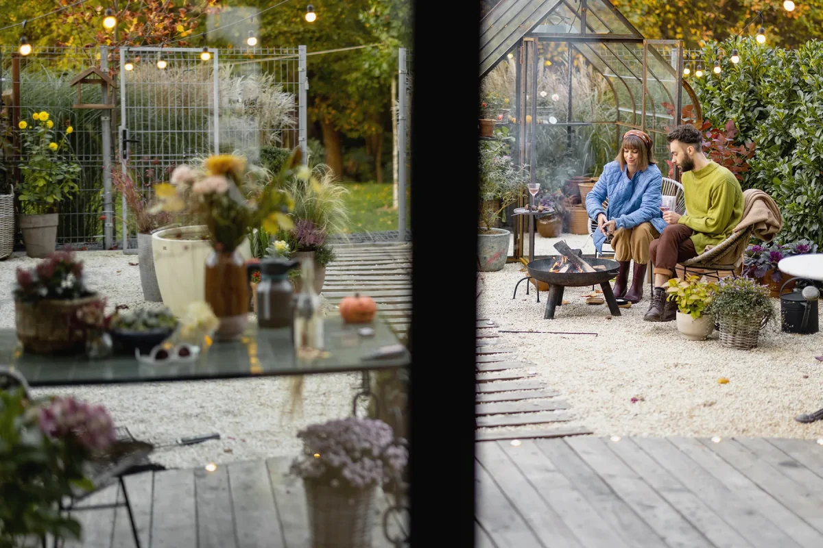 A cozy outdoor scene with a young couple sitting by a fire pit, enjoying a relaxed evening. The couple, dressed in casual and warm clothing, sits on wicker chairs in a gravel area with potted plants and a greenhouse in the background. The foreground features a glass table with various items, including a vase of flowers, and the area is lit with string lights, creating a warm and inviting atmosphere.